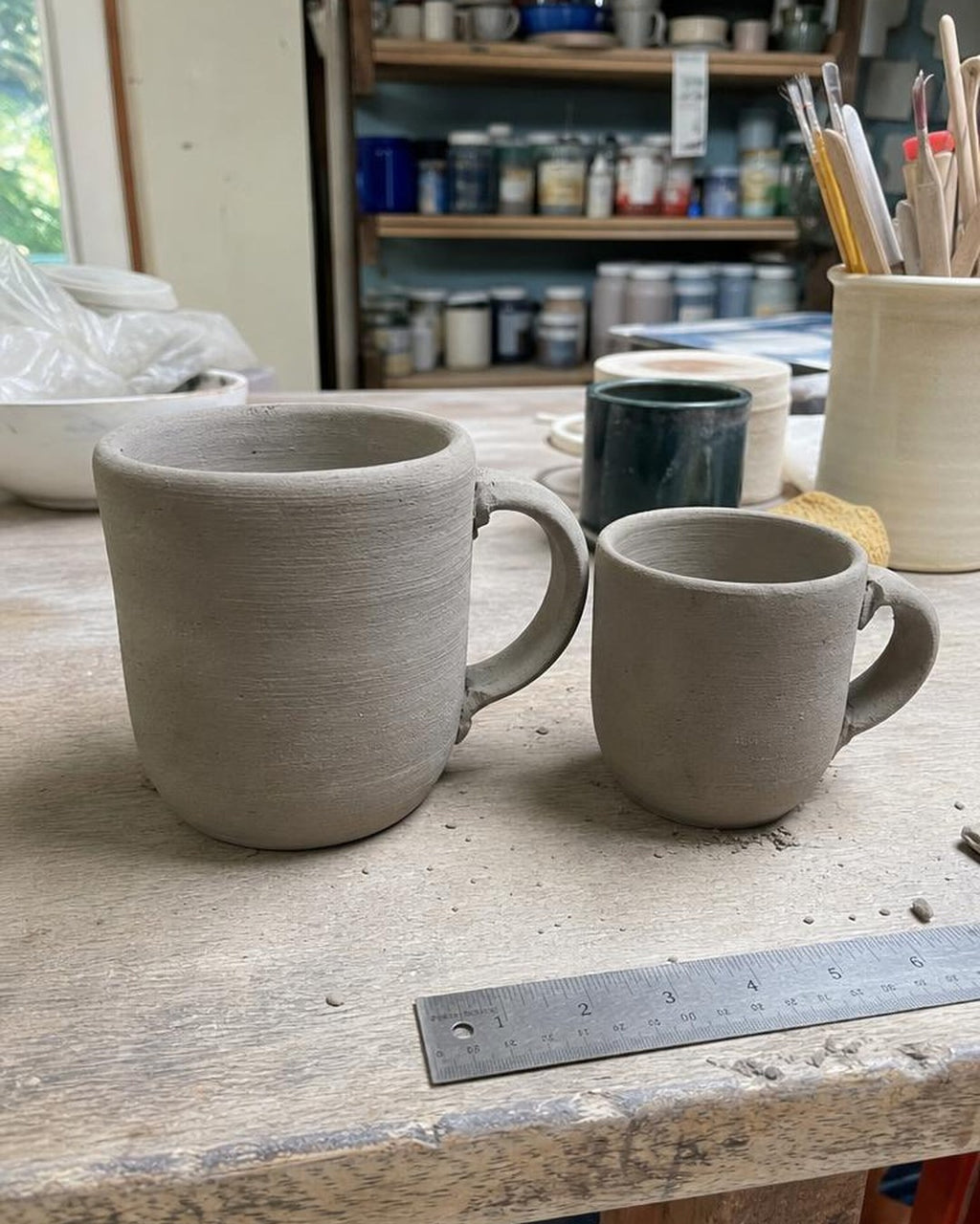Two greenware mugs side by side on a studio bench with a steel ruler